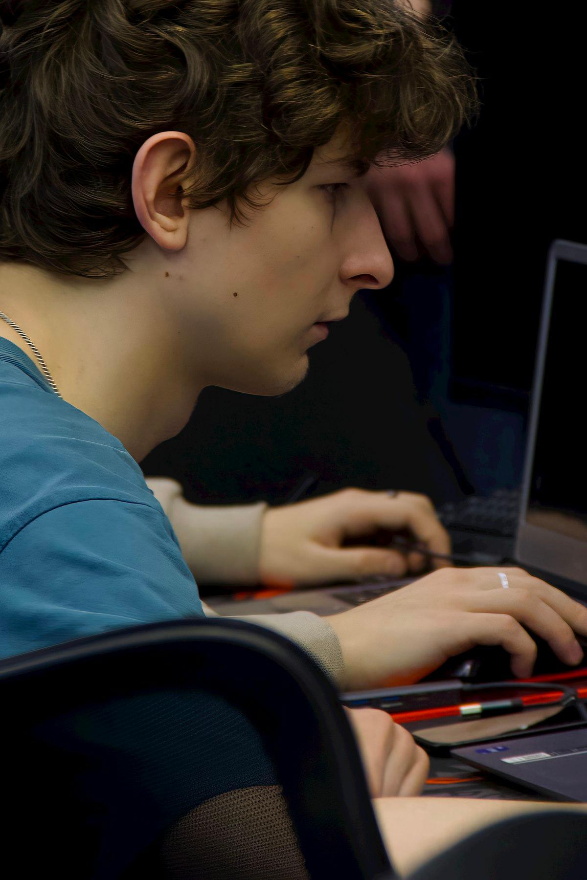 Estudiante de secundaria de perfil, con el rostro iluminado por la pantalla, trabajando en una computadora portátil dentro de un aula a oscuras